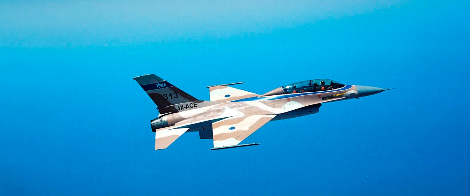 A camouflage-painted fighter jet featuring F-16 Aerostructures and Israeli Air Force markings soars against a clear blue sky, with two pilots visible in the cockpit.