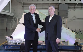 Two men in suits shake hands and smile in front of a large aircraft part inside a spacious, industrial-looking hangar.