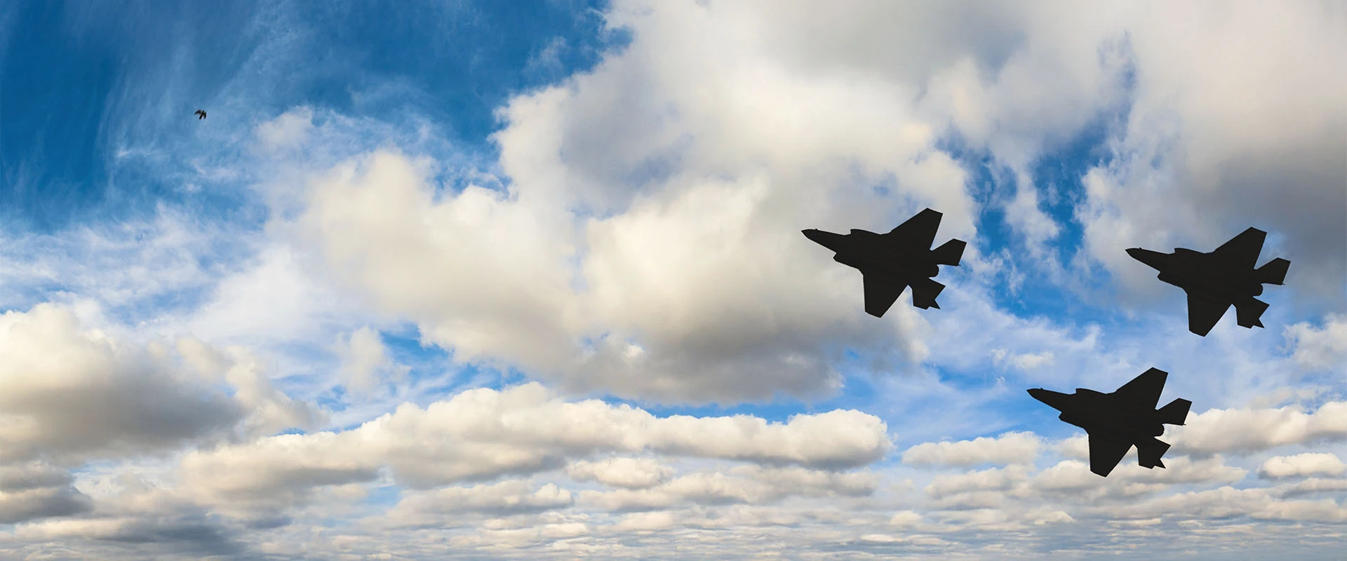 Three fighter jets featuring advanced F-35 aerostructures fly in formation against a partly cloudy blue sky, with sunlight illuminating the white clouds in the background.
