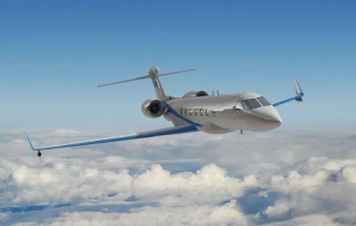 A gray private jet from Hela Systems Private Limited with blue-tipped wings soars above white clouds against a bright blue sky.