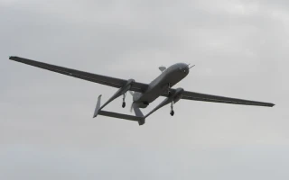 A gray unmanned aerial vehicle (drone) with long wings is flying against a cloudy sky, viewed from below.