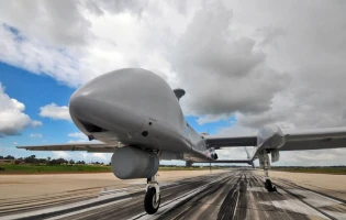 A large unmanned aerial drone equipped with advanced airborne electronics sits on a runway under a cloudy sky, with patches of blue in the background. The grey drone faces forward, highlighting its cutting-edge SIGINT Israel capabilities.