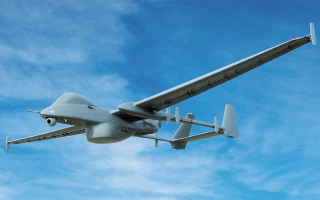 A gray military drone with long wings is flying in a clear blue sky, viewed from below and behind. The drone has cameras and sensors attached to its body.