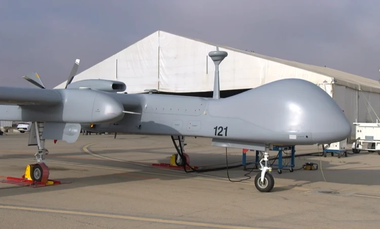 A large gray military drone, marked with the number 121, is parked on the ground in front of a white tented hangar on a cloudy day. The unmanned aerial systems operator has secured it with red and yellow wheel chocks.