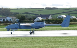 A large, gray combat unmanned aerial vehicle (UAV) is taxiing on a runway with grass on either side in India. Houses and green fields are visible in the background under a partly cloudy sky.