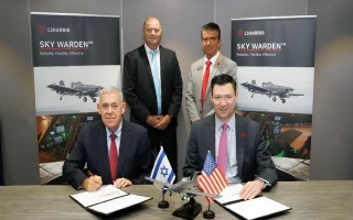 Two men sit at a table signing documents, with small Israeli and American flags and a model aircraft in front of them. Three other men stand behind. Two SKY WARDEN banners featuring IAI aviation are in the background.
