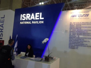 A woman sits at a reception desk in front of a blue and white booth labeled ISRAEL NATIONAL PAVILION, showcasing Maritime Solutions with a list of companies displayed on the wall and an orchid plant on the desk.