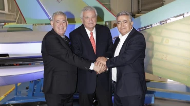 Three men in suits stand indoors, smiling and shaking hands in front of large F-35 aircraft parts or industrial equipment.