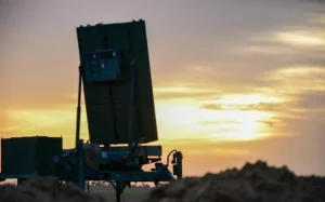 A large MMR military radar system is positioned outdoors, silhouetted against a dramatic sunset sky, with a pile of dirt in the foreground—a testament to cutting-edge defence against swarm threats.