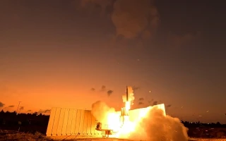 A missile launches from behind a large, white structure at dusk, bright flames and smoke illuminating the area—an impressive scene often featured in the Israel Aerospace Industries annual report.