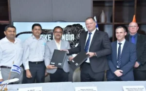 Six men in business attire stand and smile for a photo during a formal agreement signing in India. Two men in the center exchange documents, possibly related to tanker or combat initiatives, while others look on. A MAKE IN INDIA screen with a leopard graphic is behind them.
