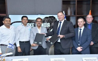 Six men in business attire stand and smile for a photo during a formal agreement signing in India. Two men in the center exchange documents, possibly related to tanker or combat initiatives, while others look on. A MAKE IN INDIA screen with a leopard graphic is behind them.