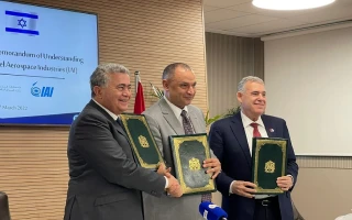 Three men in suits stand together smiling, each holding a green folder. A screen behind them shows the Israeli flag and text about a memorandum of understanding with the Morocco aerospace industry. A microphone is visible in the foreground.