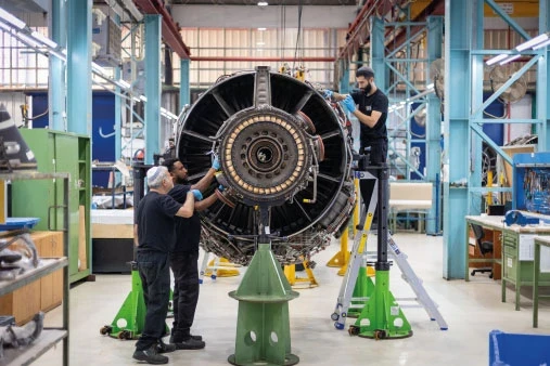Three people work on a large IAI jet engine in a brightly lit industrial workshop, using tools and ladders, surrounded by machinery and metal beams.