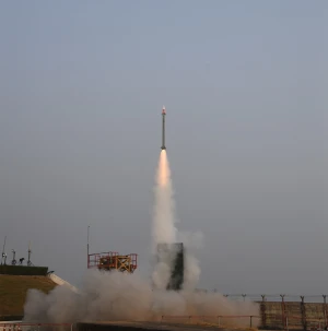 A missile launches vertically into the sky, leaving a trail of smoke behind it, with a clear sky and some structures visible on the ground below.