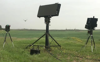 Three electronic devices with large screens and stands are set up in a grassy field. A small drone is visible flying in the distance against a cloudy sky. Cables connect the devices on the ground.