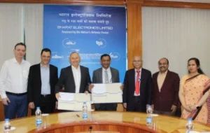 Seven people stand in front of a blue Bharat Electronics Limited banner, holding documents and smiling for a group photo after an ADSN BEL Israel event in a conference room with a round table and water bottles.