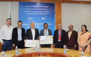 Seven people stand in front of a blue Bharat Electronics Limited banner, holding documents and smiling for a group photo after an ADSN BEL Israel event in a conference room with a round table and water bottles.