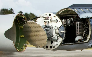Close-up of an aircraft’s nose with the radar system exposed, showing internal electronic components and wiring. The surrounding panels are open, revealing the radar array and internal structure.