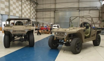Two tan, military-style combat vehicles are parked inside a large hangar. The vehicles have rugged tires and minimal frames. People and other vehicles are visible in the background.