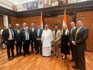 A group of people in formal attire stands in a wood-paneled room with two Indian flags, a large emblem, and portraits on the wall behind them. The central figure is dressed in traditional Indian clothing.