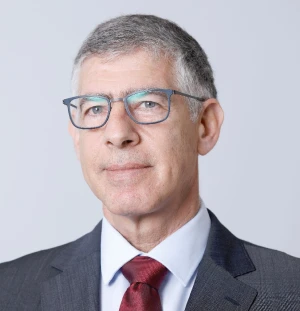 A middle-aged man with short gray hair and glasses, wearing a dark suit, white shirt, and red tie, poses against a plain light gray background.