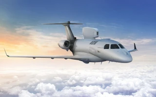 A gray military surveillance aircraft flies above white clouds with a blue and orange sky in the background.