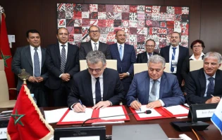 A group of men and women in business attire pose for a photo as two men seated at a table sign documents, representing the International University of Rabat. Red and white abstract artwork and the flag of Morocco are visible in the background.