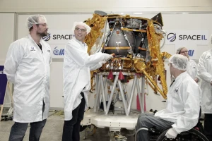 Several people in white lab coats and hairnets stand around and examine a partially assembled spacecraft in a laboratory, with SpaceIL logos visible in the background, as they carefully inspect a time capsule destined for space.
