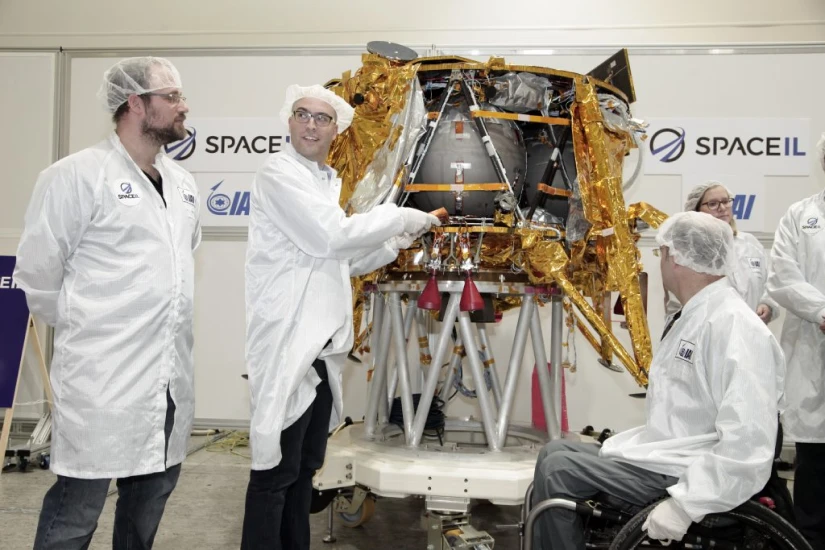 Four scientists in white lab coats and hairnets stand near a gold and silver lunar lander at a SpaceIL facility, discussing its features. One points toward the lander’s time capsule, while another sits in a wheelchair.