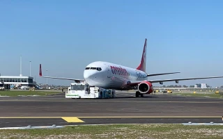 A large white and red passenger airplane is being towed on an airport runway under a clear blue sky. Airport buildings and grassy areas are visible in the background.