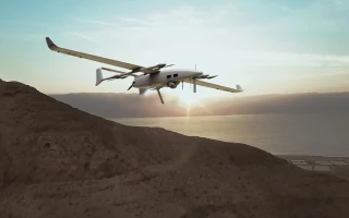 A white drone with twin propellers flies over a rocky hill at sunset, with a body of water and distant mountains visible in the background.