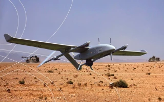 A military drone utilizing VTOL technologies flies low over a desert landscape with sparse vegetation; two military vehicles are visible in the background under a clear sky.