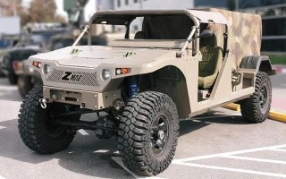 A tan, rugged military-style off-road vehicle with large tires and a camouflage-patterned rear cover is parked on a city street.