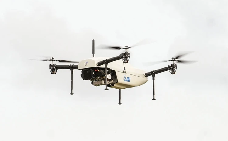 An IAI white quadcopter drone with four propellers and landing legs is flying against a cloudy sky.