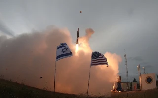 A rocket launches into the sky, surrounded by thick smoke, with Israeli and American flags in the foreground waving in the wind.