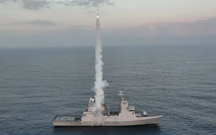 A warship at sea launches an IAI missile vertically, leaving a trail of white smoke rising into the sky. The ocean and a cloudy sky are visible in the background.
