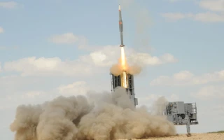 An IAI missile is being launched from a ground-based launcher, with flames and smoke billowing out at the base and clouds in the sky above.