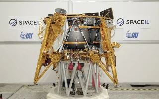 A spacecraft covered in gold foil stands indoors on a support structure. SpaceIL and IAI logos are visible on the white wall behind it.
