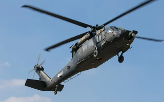 A black military helicopter with spinning rotor blades is flying in a clear blue sky, viewed from below and slightly to the side.