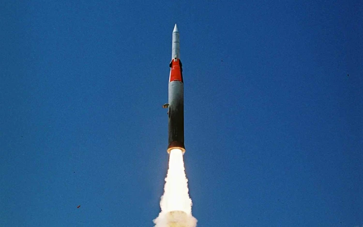 A rocket from IAI launches vertically into a clear blue sky, emitting bright flames and white smoke from its engines. The rocket is mostly gray with a red and white section near the top.