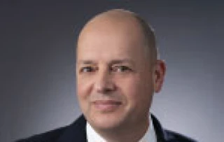 A middle-aged man with a shaved head, wearing a dark suit and white shirt, is posing for a professional headshot against a plain, dark background.