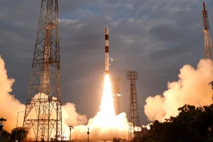 A rocket launches into the sky, surrounded by billowing smoke and bright flames, with tall metal towers on either side and a cloudy sky in the background.