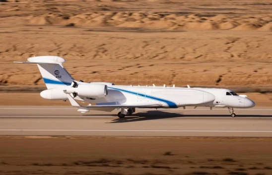 A white and blue multi mission surveillance aircraft with an enlarged radar dome on the tail is taxiing on a runway in a desert landscape.