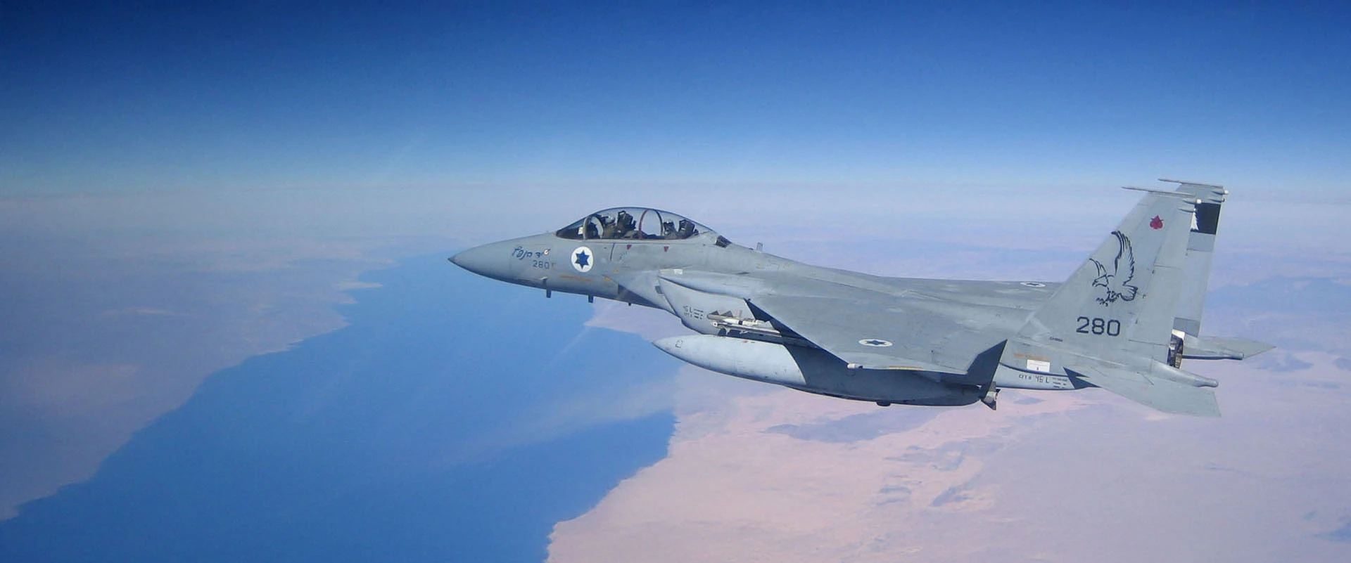 A gray military fighter jet featuring F-15 aerostructures and Israeli Air Force markings flies above a coastline, with desert and blue sea visible below under a clear sky.