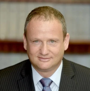 A man with short light brown hair wearing a dark pinstripe suit, light blue shirt, and patterned tie, smiling slightly, with blurred bookshelves in the background.