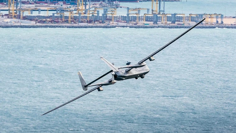 An IAI military drone with wide wings flies over the ocean near a busy industrial port, cranes, and shipping containers in the background.