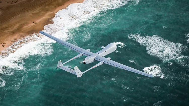 An IAI unmanned aerial vehicle (drone) with long wings glides over turquoise ocean waves near a sandy coastline.