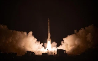 A rocket launches into the night sky, carrying a satellite amid billowing clouds of smoke and illuminated by fiery engine light, with dark silhouettes of structures visible in the foreground.