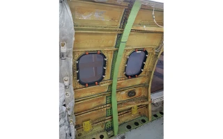 Interior view of an aircraft fuselage showing two window openings with no window panes, exposed metal framework, visible structural supports, rivets, and pickle fork frame fittings.